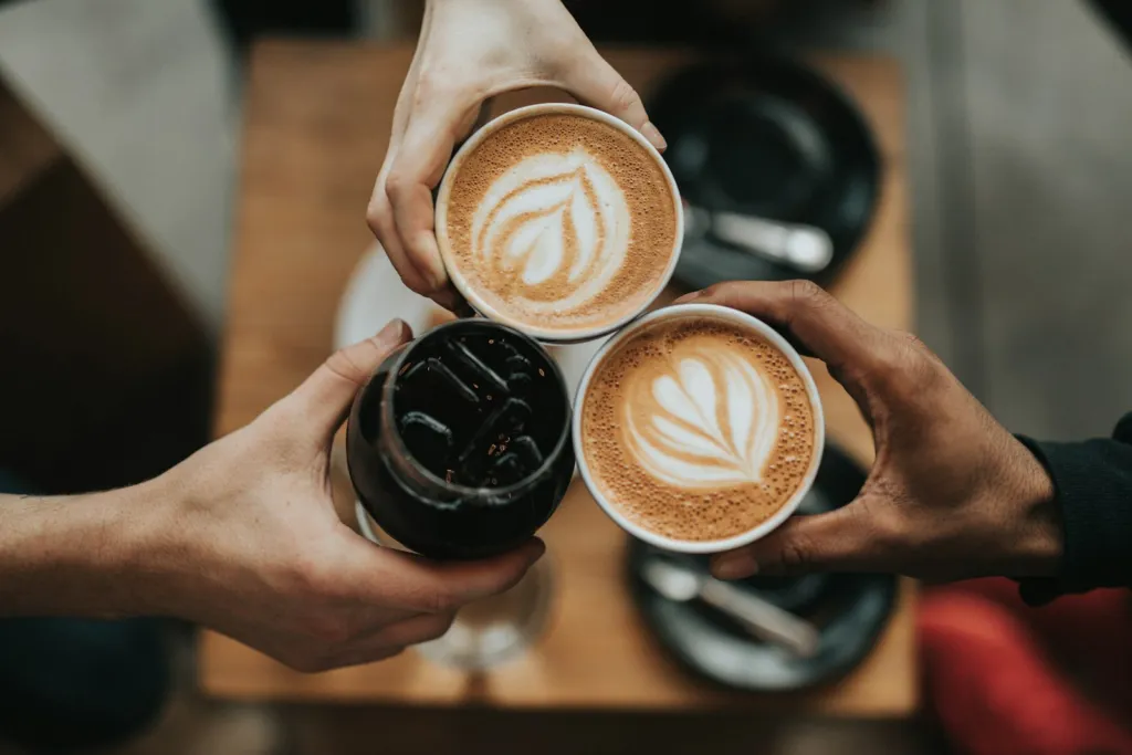 Three hands clinking cups of latte and coffee together over a wooden table